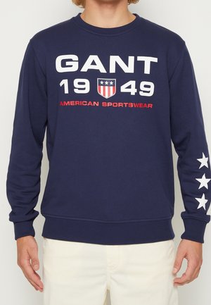 Person wearing navy sweatshirt with white text "GANT 1949" and shield logo, red "American Sportswear," and white stars down right sleeve.