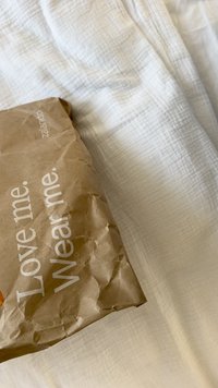 Barna papírdoboz gyűrött textúrával, fehér felirattal, amely a következőt mondja: "Szeress engem. Viselj engem." A logó látható az oldalon, fehér szöveten pihen.