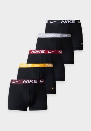 Νέος άνδρας στέκεται ξυπόλητος με μαύρα boxer Nike μπροστά σε απλό φωτεινό φόντο, με το ένα χέρι πίσω από την πλάτη του.