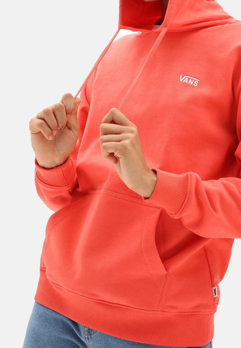 Personne portant un sweat à capuche Vans corail vif tenant les cordons de la capuche, associé à un jean bleu.