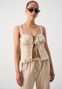 Crop top en lin beige avec décolleté carré, lien frontal ajustable et larges bretelles ; associé à un pantalon beige large avec cordon de serrage.