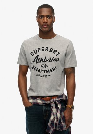 Grijze katoenen t-shirt met een zwarte grafische bedrukking die zegt "SUPERDRY Athletics Department." Gecombineerd met donkere jeans en een geruite overhemd om de taille geknoopt.