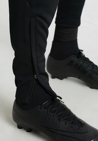 Schwarze Sporthosen mit Reißverschlüssen an den Seiten, die eine glatte Textur aufweisen. Kombiniert mit schwarzen Fußballschuhen mit strukturierten Mustern.
