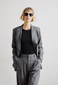Grå mönstrad blazer över en svart topp, tillsammans med ljusgrå högmidjade byxor. Modellen bär runda svarta solglasögon.