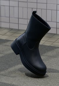 Μαύρο δερμάτινο μποτάκι αστραγάλου με χοντρή σόλα, στέκεται όρθιο πάνω σε υφή πεζοδρομίου μπροστά σε φόντο από πλακάκια.