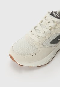 Light beige sneaker crafted from mesh and suede, featuring a textured toe, laces, a brown rubber outsole, and a logo on the tongue.