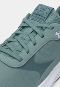 Tuvinājums uz tumši zaļo Under Armour Aurora kedu, uzsvaru liekot uz strukturēto sietu, auklām un zīmola logotipa detaļām uz mēles.