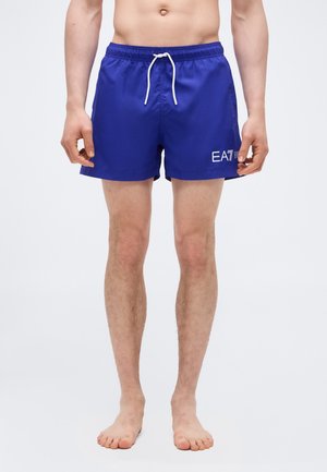 Άνδρας με μπλε μαγιό σορτς με λευκό κορδόνι και λευκό λογότυπο "EA7", στέκεται ξυπόλυτος μπροστά σε απλό φόντο.
