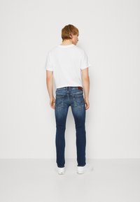 Vaqueros ajustados de un azul oscuro con un diseño desgastado, cinco bolsillos y un parche de cuero marrón en la parte trasera de la cintura. Combinados con una camiseta blanca sencilla.