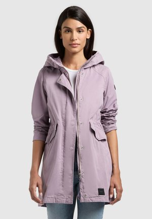 Femme portant une veste à capuche violet clair avec des poches avant sur un haut blanc et un jean bleu, debout devant un fond uni.