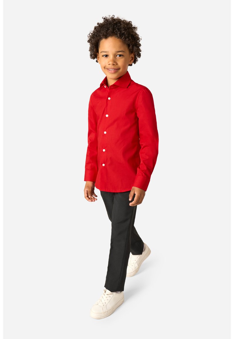 Junge, der nach vorne geht und ein leuchtend rotes Hemd mit Knöpfen, schwarze Hosen und weiße Turnschuhe trägt, vor einem schlichten weißen Hintergrund.