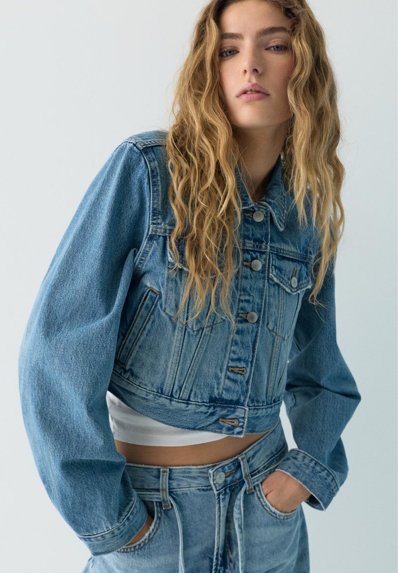 Jeune femme aux longs cheveux blonds ondulés portant une veste en jean bleue courte et un jean taille haute assorti, les mains dans les poches.
