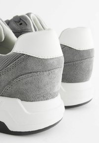 Grå sneakers har en design i mocka och nät med en vit läderaccent på hälen och en texturerad gummisula. Närbild på baksidan.