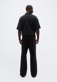 Ein Mann steht mit dem Rücken zur Kamera, trägt lockere schwarze Hosen und ein schwarzes kurzärmeliges Hemd mit Kragen vor einem schlichten hellen Hintergrund.