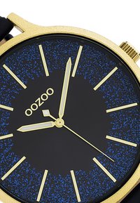 Montre dorée avec un cadran noir arborant des accents bleu scintillant. Elle possède des index et des aiguilles dorées, ainsi qu'un boîtier rond avec un bracelet noir.