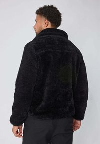 Homme aux cheveux bouclés courts portant une veste noire en peluche et un pantalon noir, se tenant de dos contre un fond clair uni.
