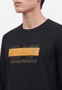 Junge Person trägt ein schwarzes Langarmshirt mit dem "EA7 Emporio Armani"-Logo in Gold und Orange auf der Brust.