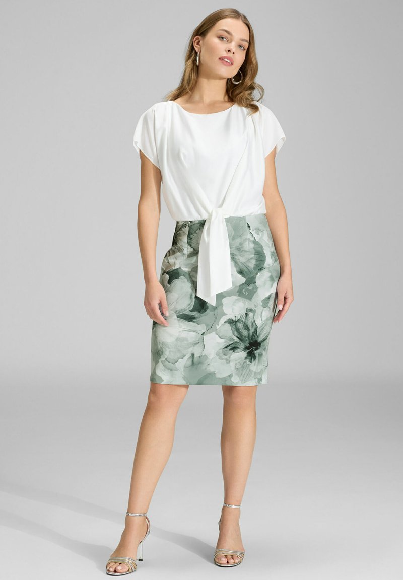 Blouse blanche nouée à la taille, associée à une jupe crayon longueur genou à imprimé floral vert. Le mannequin porte des talons à brides argentées.