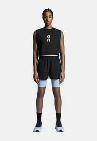 Schwarzes ärmelloses Crop-Top mit Logo, kombiniert mit schwarzen Shorts und hellblauen Leggings. Das Modell trägt schwarze Socken und graue Sportschuhe.
