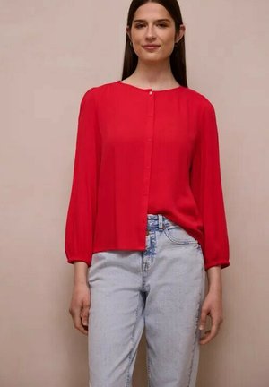 Kvinde iført en rød langærmet skjorte med knapper og lyseblå højtaljede jeans, stående foran en ensartet beige baggrund.