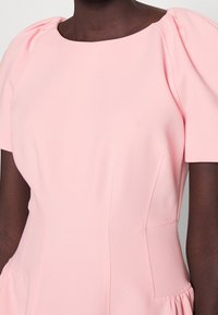 Hellrosa Kleid mit kurzen Puffärmeln, Rundhalsausschnitt, seitlichen Taschen und gerafften Details in der Taille aus einem glatten Stoff.