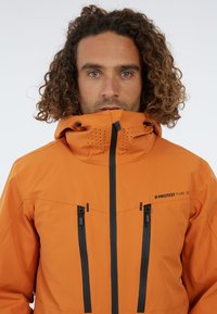 Chaqueta impermeable naranja con un alto cuello, bolsillos con cremallera y detalles en negro. El material es suave con un acabado mate.