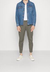 Veste en denim avec deux poches poitrine, couleur bleu clair, et fermeture à boutons. Pantalons vert olive fuselés et baskets blanches avec des œillets multicolores.