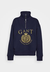 Sweat-shirt bleu marine à fermeture quart de zip avec col haut, logo jaune GANT, blason couronné et texte "American Sportswear" sur le devant.
