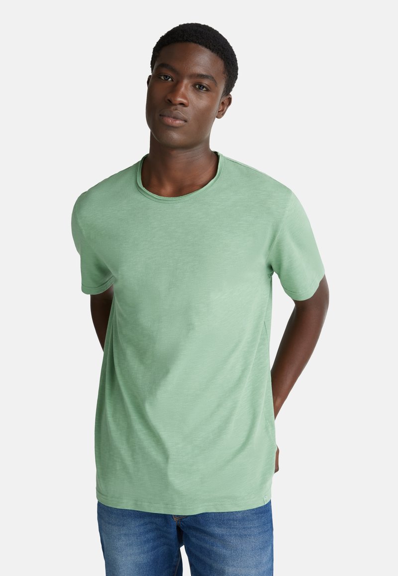 T-shirt verde a maniche corte realizzato in tessuto morbido con scollatura rotonda, abbinato a jeans blu. La maglietta presenta una consistenza sottile.