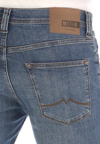 I jeans in denim blu presentano una tasca posteriore con un design distintivo e una toppa in pelle marrone etichettata "MUSTANG True Denim." Dettagli cuciti presenti.