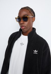 Zwarte fleece-jas met hoge kraag, witte logo op de borst, gelaagd over een wit T-shirt. Geelgetinte vierkante zonnebril.