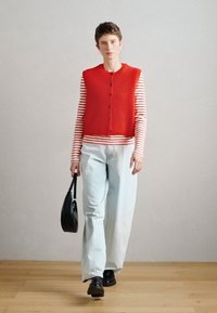 Κόκκινο πλεκτό γιλέκο με ραβδώσεις και κουμπιά, φορεμένο πάνω από ένα ριγέ μακρυμάνικο πουκάμισο. Ανοιχτό γαλάζιο παντελόνι με φαρδιά γραμμή και μαύρα παπούτσια. Μαύρη τσάντα χειρός.