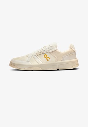 Baskets basses blanc cassé avec panneaux en cuir et daim, semelle beige, fermeture à lacets et logo jaune « OC » sur le côté.