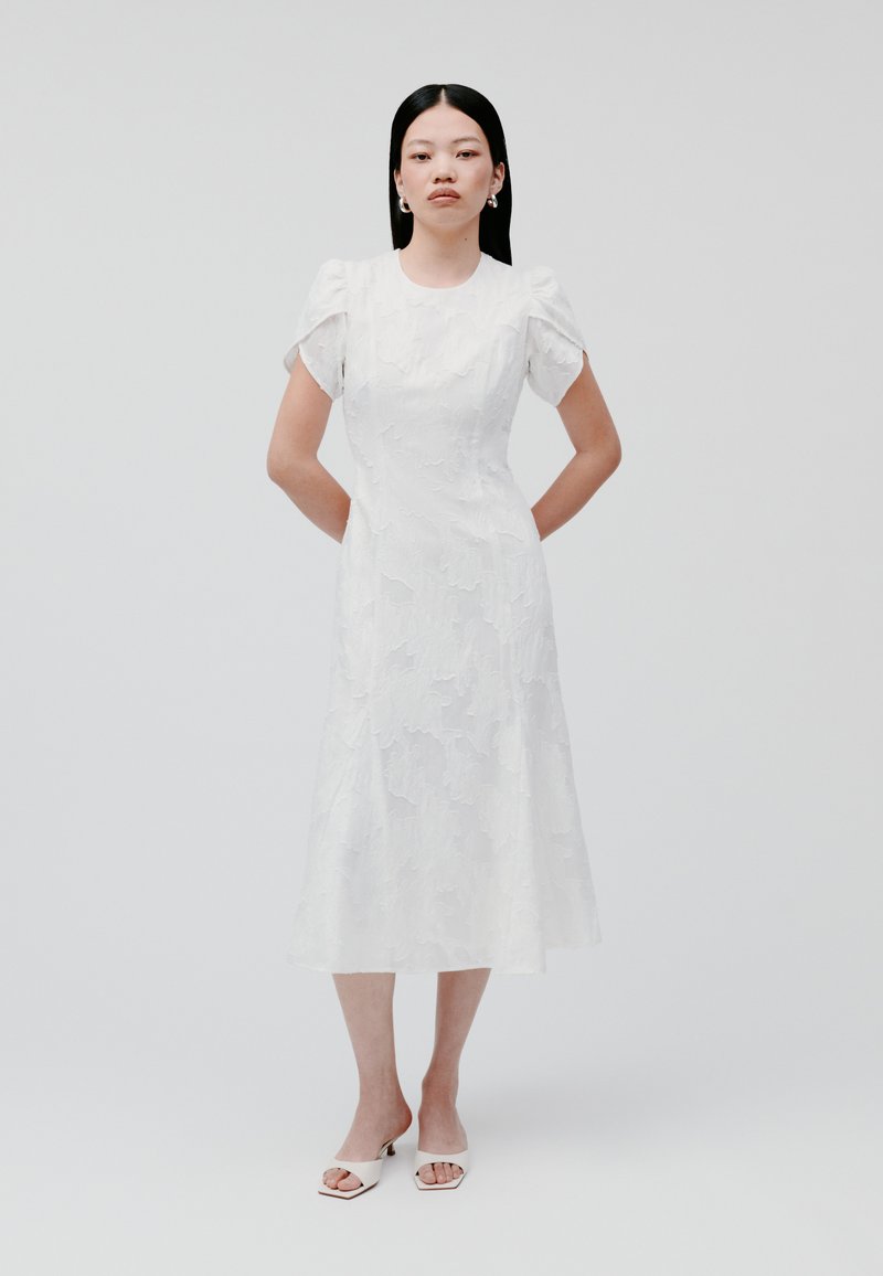 Abito midi bianco con maniche a palloncino, motivo floreale testurizzato, corsetto aderente e gonna svasata. Il modello indossa tacchi bianchi aperti.