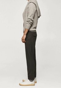 Lichtgrijze gebreide trui over de schouders gedrapeerd, gecombineerd met donkergroene broek en witte sneakers met een beige zool. Zijaanzicht.