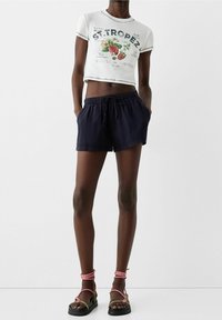 Bílý crop top s grafickým potiskem, námořnické šortky s elastickým pasem a sandály na řemínky s růžovými akcenty a černými podrážkami.