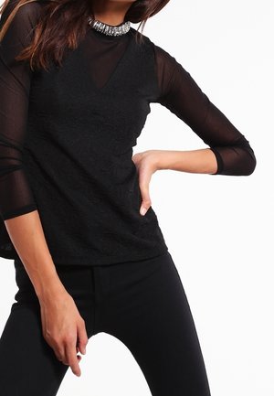 Frau trägt ein schwarzes, durchsichtiges Langarm-Top mit einem mit Schmucksteinen besetzten Kragen und schwarze Hosen, Hand in der Hüfte, posiert vor weißem Hintergrund.