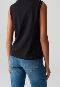 Schwarzes ärmelloses Oberteil aus Baumwolle mit rundem Halsausschnitt und lockerer Passform. Kombiniert mit hellblauen Jeans mit Gesäßtaschen.