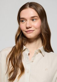 Beżowa koszula z guzikami, wykonana z lekkiego materiału, z małym logo na lewej piersi i klasycznym wzorem kołnierzyka.