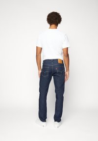 Jeans de mezclilla azul oscuro con pierna recta, con un parche de cuero en la parte posterior, costuras amarillas y bolsillos traseros. Llevados con una camiseta blanca.