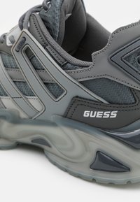Baskets grises avec tige tissée, accents brillants et semelle translucide épaisse. Logo "GUESS" affiché sur le côté. Design texturé et aérodynamique.