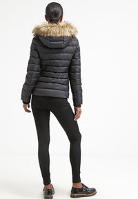 Černá péřová bunda s umělým kožešinovým kapucí, žebrovanými manžetami a prošívanou texturou. Nosič má na sobě černé slim-fit kalhoty a ploché boty.