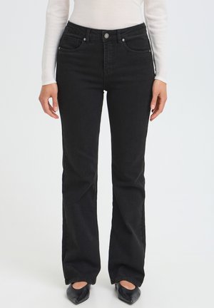 Femme portant un jean noir droit, un haut blanc à manches longues et des chaussures noires à bout pointu, debout sur un fond uni.