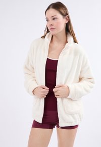 Krémová fleecová bunda s vysokým límcem, zapínáním na zip a bočními kapsami, nošená přes bordó tílko a šortky. Hladká textura.