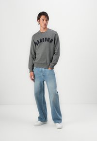 Γκρι φούτερ με το κείμενο "BARBOUR" σε ναυαρινό, συνδυασμένο με ανοιχτό μπλε παντελόνι σε φαρδιά γραμμή και λευκά αθλητικά παπούτσια σε ουδέτερο φόντο.