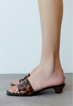 Persoon die open teen, laaggehakte sandalen met bruin en zwarte schildpadmotiefriemen draagt, staand op een grijze vloer.
