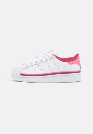 Hvid Adidas Superstar sneaker med lyserød indvendig foring, hælflig og mellemsålstribe, set fra siden på en ensfarvet baggrund.