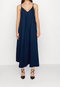 Mujer vestida con un vestido midi sin mangas de color azul marino con tirantes finos y tacones negros de punta con correas en los tobillos, de pie sobre un fondo blanco.