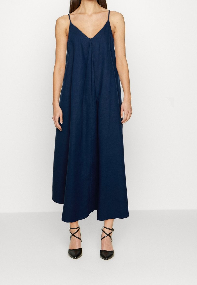 Mujer vestida con un vestido midi sin mangas de color azul marino con tirantes finos y tacones negros de punta con correas en los tobillos, de pie sobre un fondo blanco.