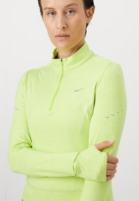 Vrouw in een neongroene lange mouw sporttop met een kwart ritssluiting en duimgaten, met de armen gekruist tegen een witte achtergrond.